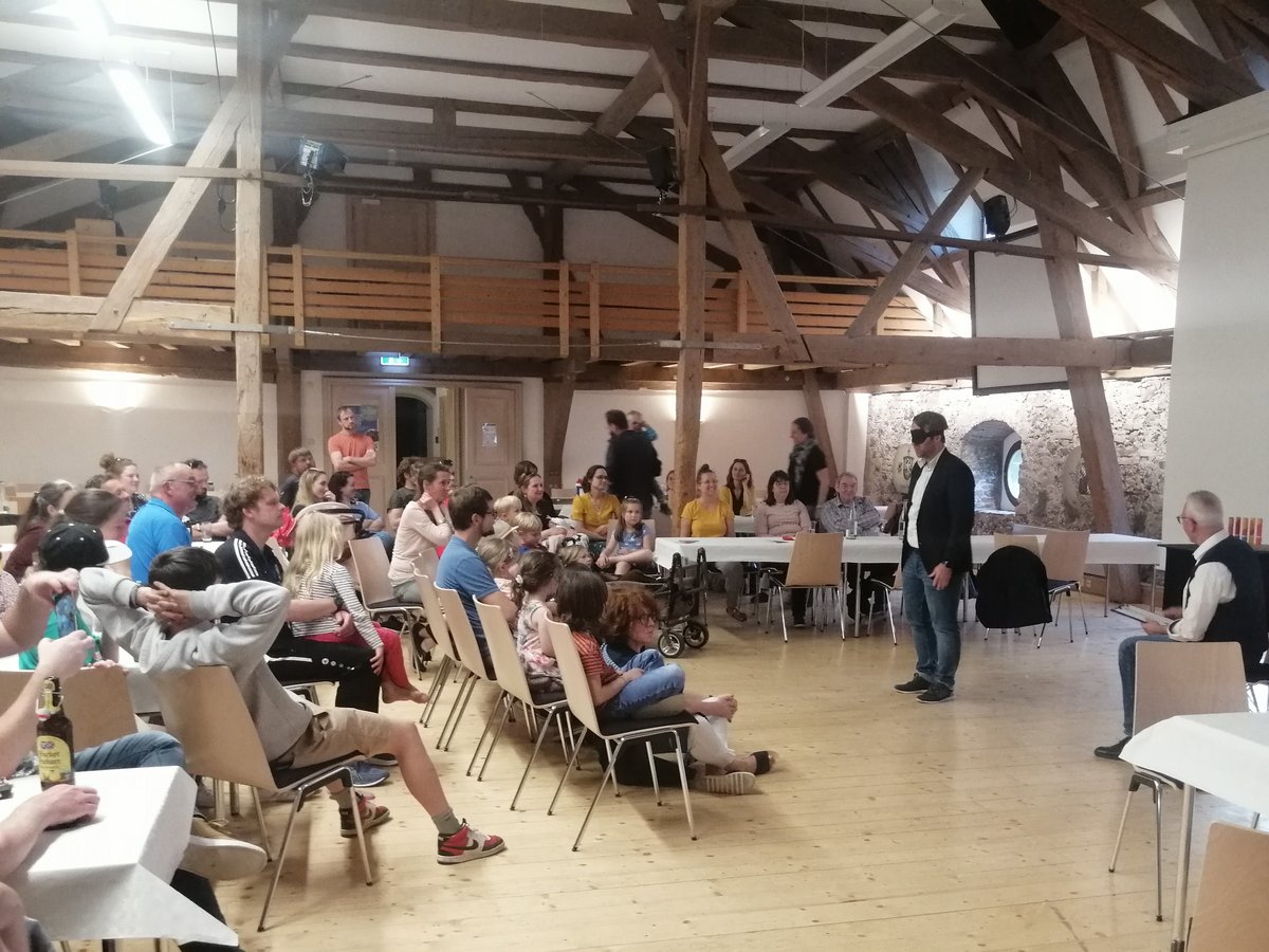 Ein Mann mit Augenbinde präsentiert vor einem Publikum aus Erwachsenen und Kindern in einem rustikalen Saal mit Holzbalken.