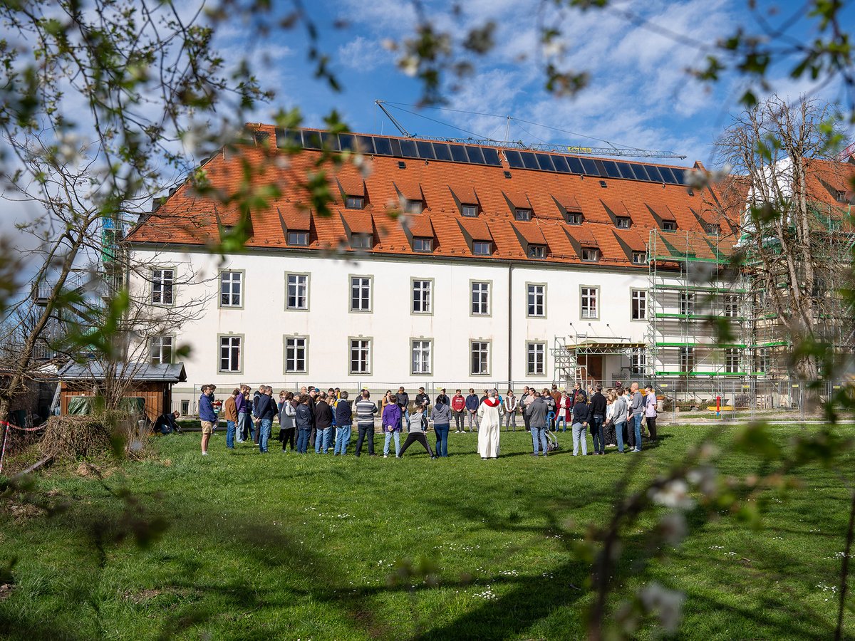 Gruppe mit gewandeter Figur auf sonnigem Rasen vor weißem Gebäude mit rotem Dach, Solarpaneelen und Baugerüst.