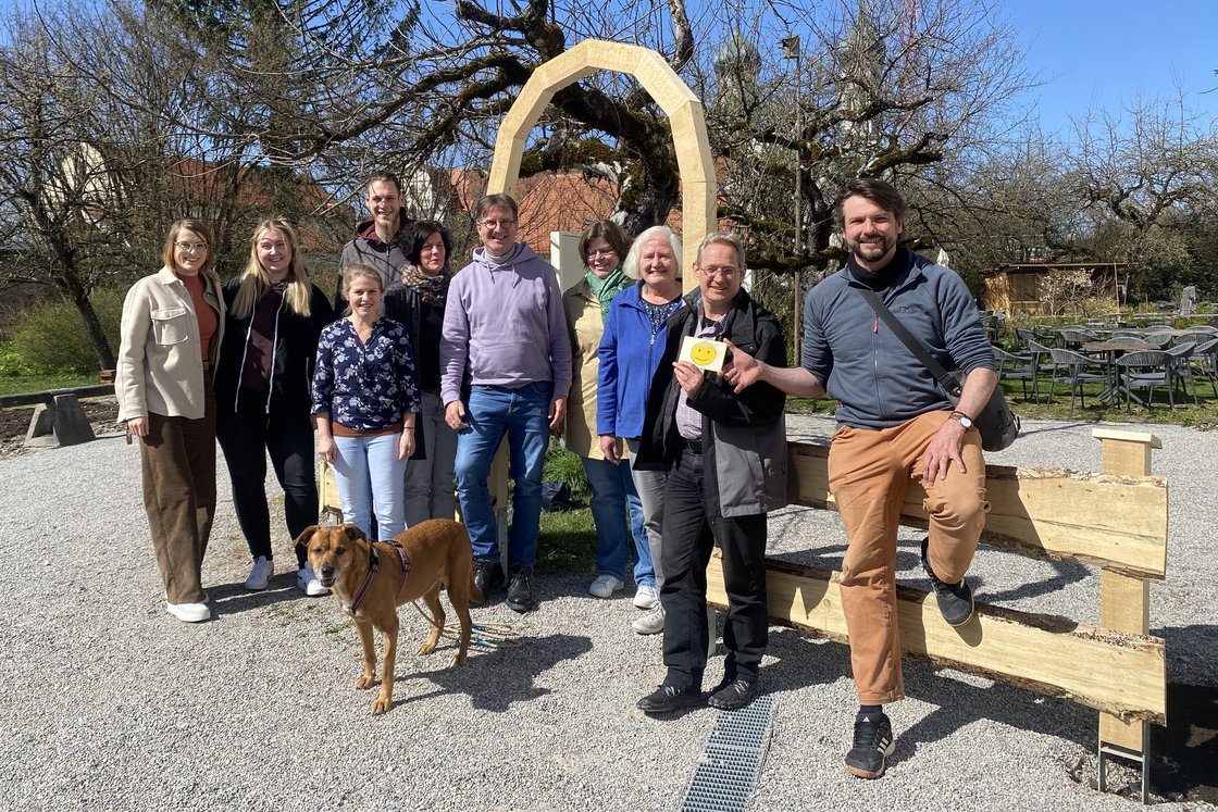 Eine lächelnde Gruppe von elf Personen und einem Hund steht im Freien vor einem Holzbogen und Bäumen an einem sonnigen Tag.