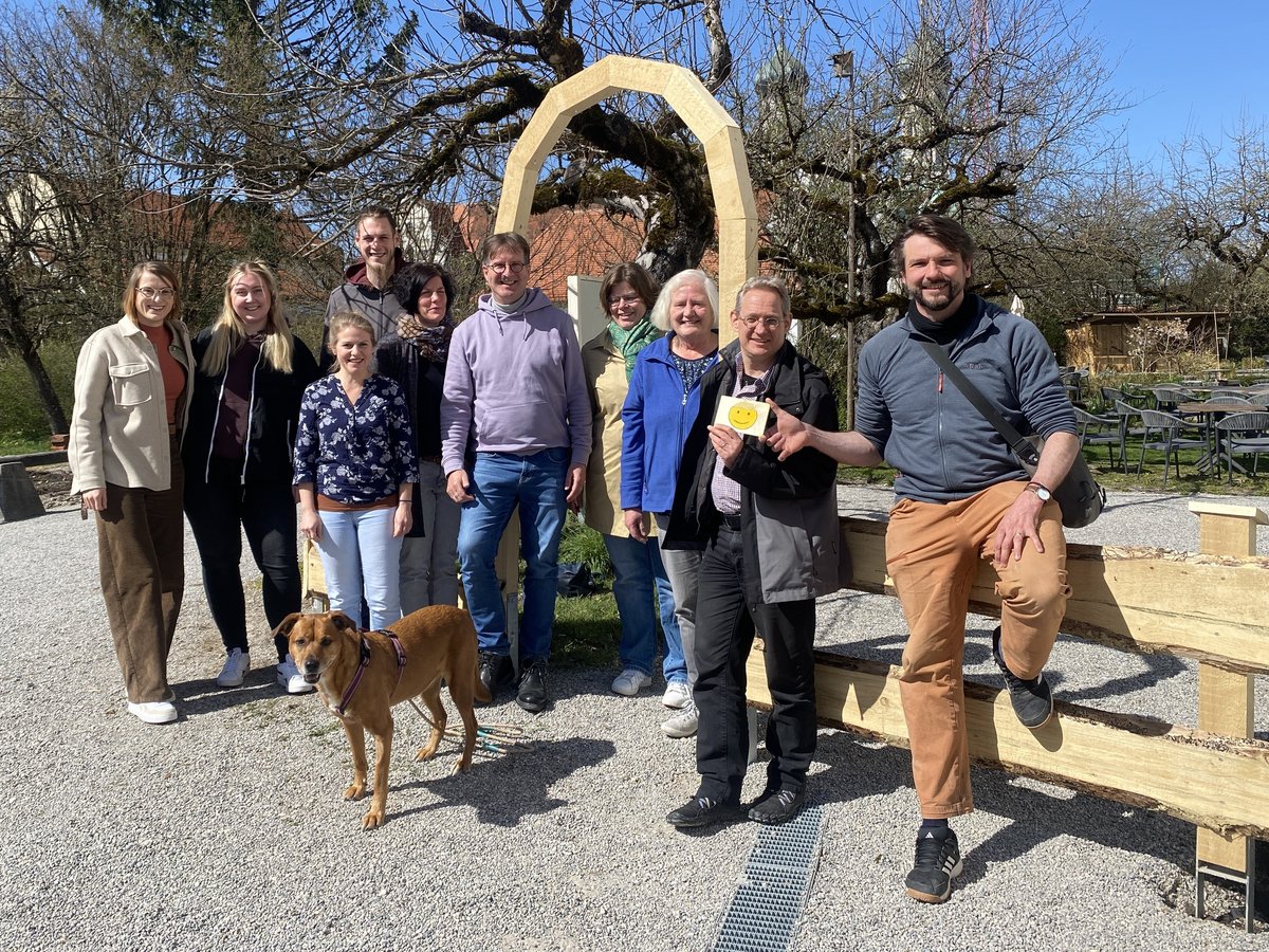 Eine lächelnde Gruppe von elf Personen und einem Hund steht im Freien vor einem Holzbogen und Bäumen an einem sonnigen Tag.