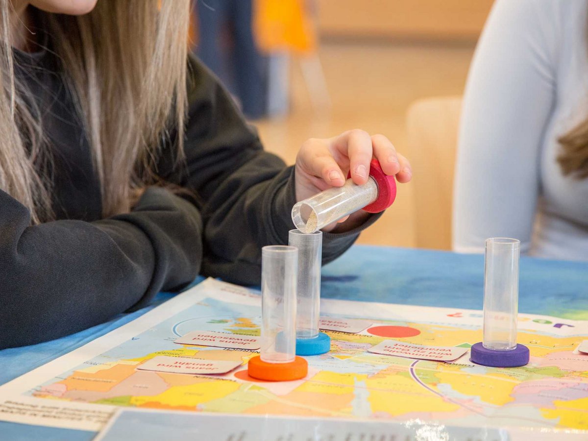 Eine Person schüttet Sand aus einem Behälter in ein Röhrchen auf einer Weltkarte. Andere Röhrchen mit farbigen Böden und Karten.
