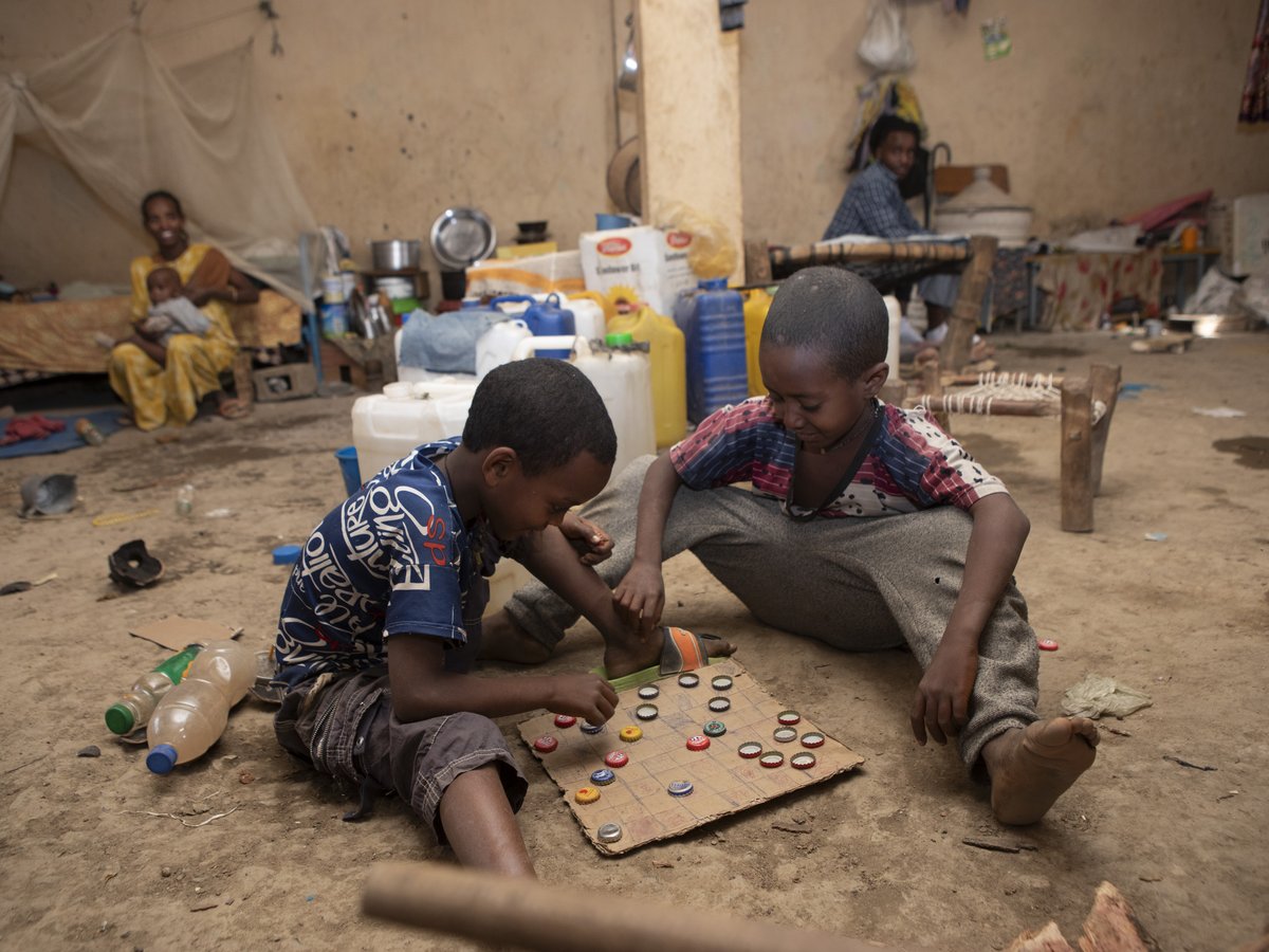 Zwei Jungen spielen ein Spiel mit Kronkorken auf einem Kartonbrett auf Lehmboden, während weitere Familienmitglieder im Hintergrund zu sehen sind.