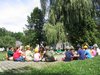 Kinder und Erwachsene sitzen im Kreis auf einem gepflasterten Platz im Freien an einem sonnigen Tag.