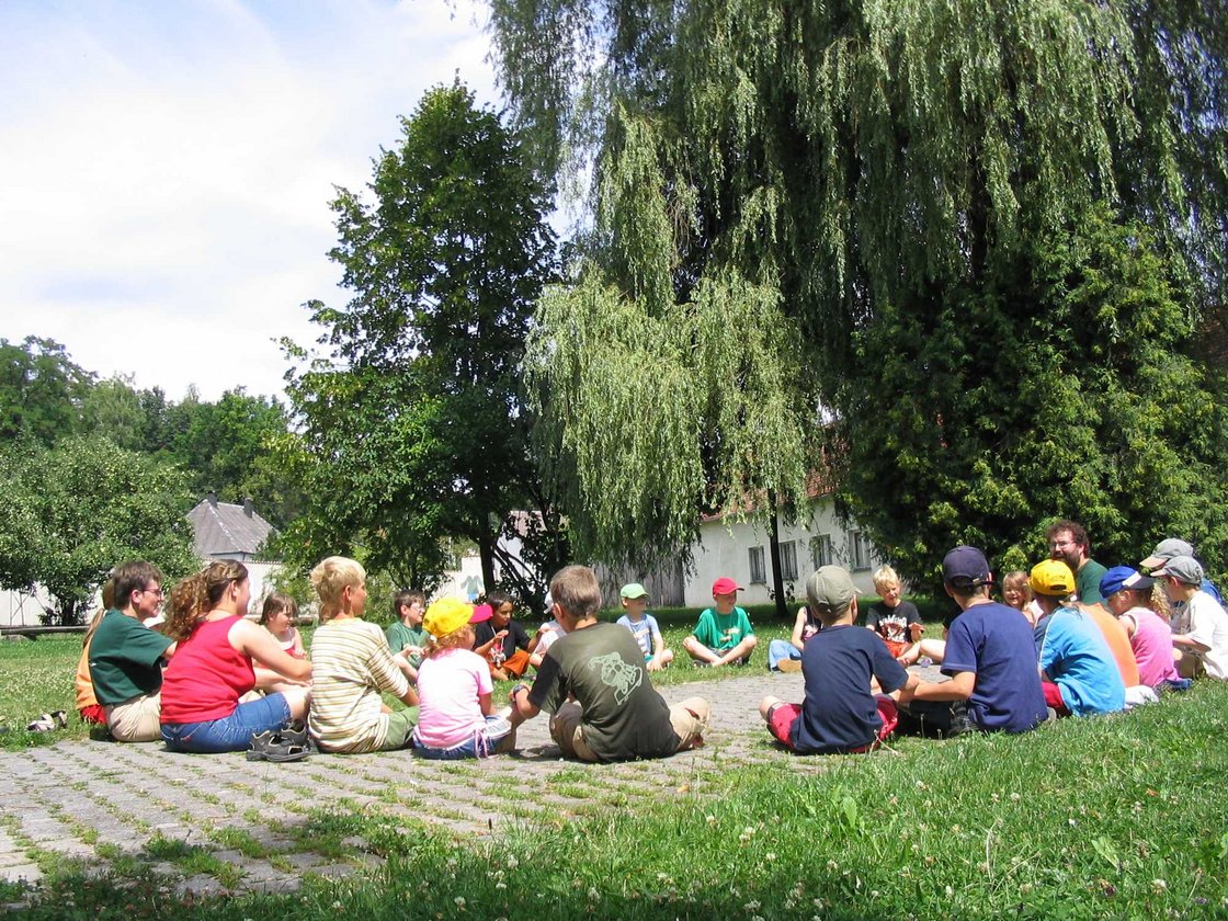 Kinder und Erwachsene sitzen im Kreis auf einem gepflasterten Platz im Freien an einem sonnigen Tag.