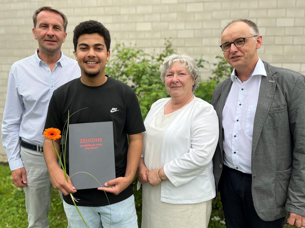 Abschlussfeier: Junger Mann hält stolz Zeugnis und orange Blume, flankiert von zwei Männern und einer Frau.