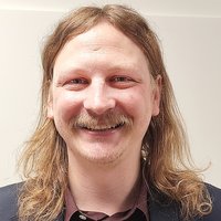 Lächelnder Mann mit langen hellbraunen Haaren, Schnurrbart und Ziegenbart, trägt eine dunkle Anzugjacke und ein weinrotes Hemd.