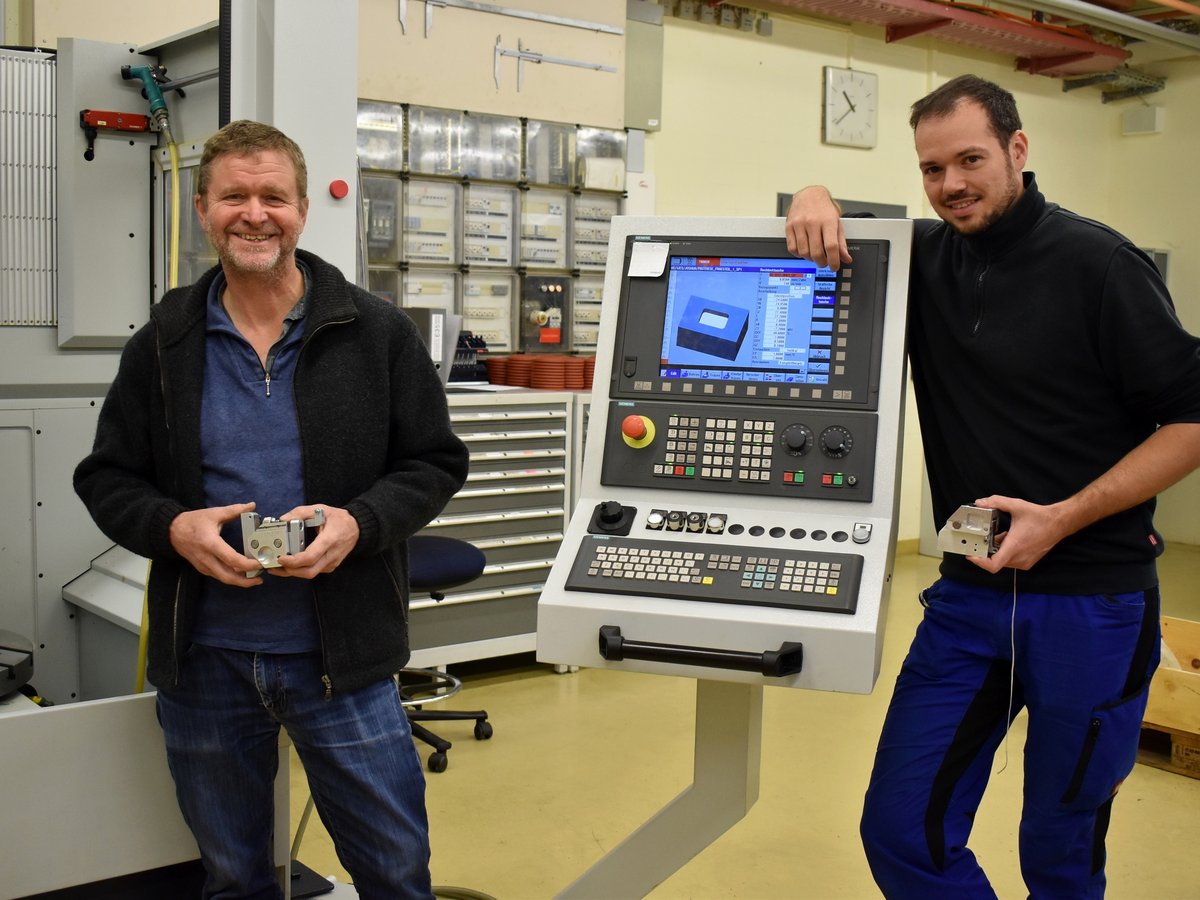 Zwei lächelnde Männer in einer Fabrik halten gefertigte Metallkomponenten neben einem CNC-Bedienfeld.