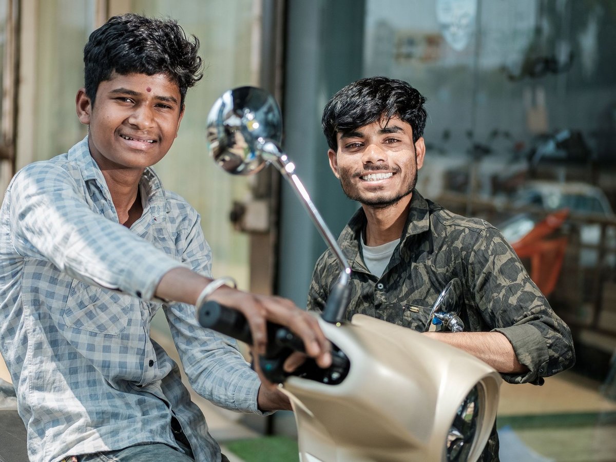 Zwei lächelnde junge Männer, einer mit Bindi, auf einem hellen Roller, glücklich in die Kamera blickend.