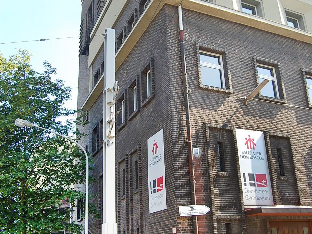 Backsteingebäude des Don-Bosco-Gymnasiums mit Schildern der Salesianer Don Boscos an einer Straße mit Baum und Laterne.