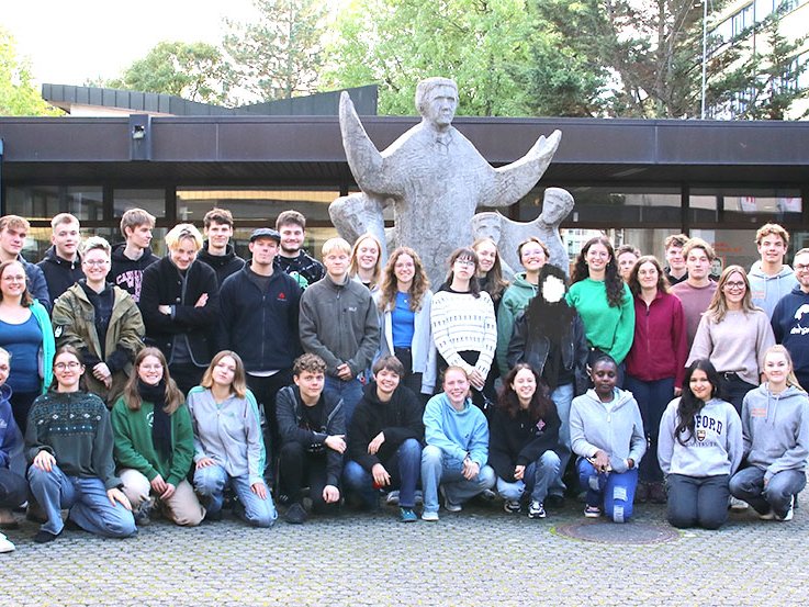 Eine vielfältige Gruppe von Studenten und Erwachsenen posiert für ein Foto im Freien vor einem modernen Gebäude und einer großen Steinskulptur.