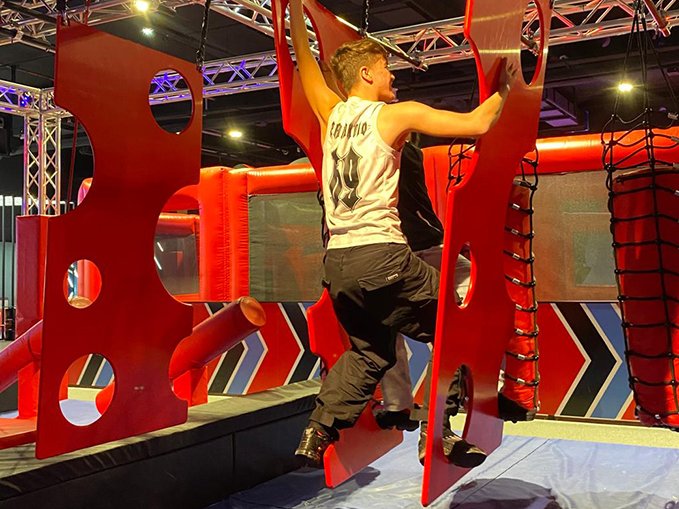 Ein junger Mann in einem weißen Tanktop und schwarzen Hosen navigiert einen Indoor-Hindernisparcours an roten Hängeelementen.