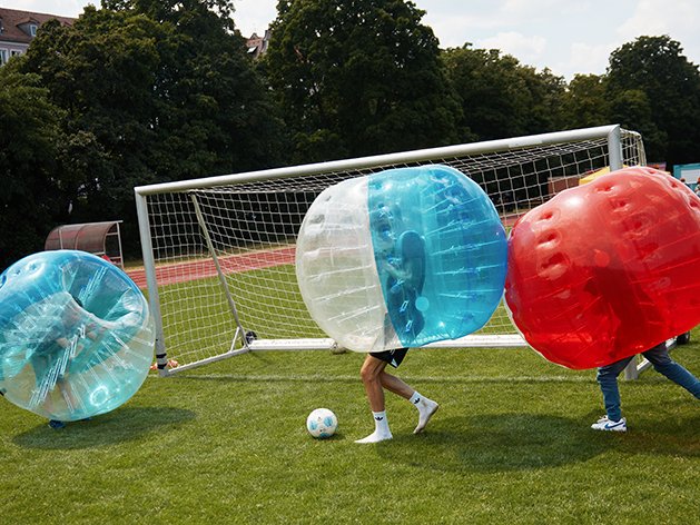 Drei Personen in blauen, weißen und roten aufblasbaren Blasenanzügen spielen Fußball auf einem Grünfeld nahe eines Tors.