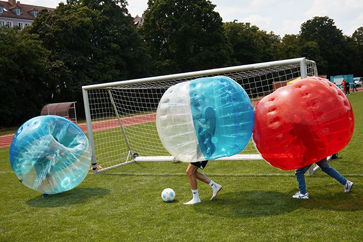Drei Personen in blauen, weißen und roten aufblasbaren Blasenanzügen spielen Fußball auf einem Grünfeld nahe eines Tors.