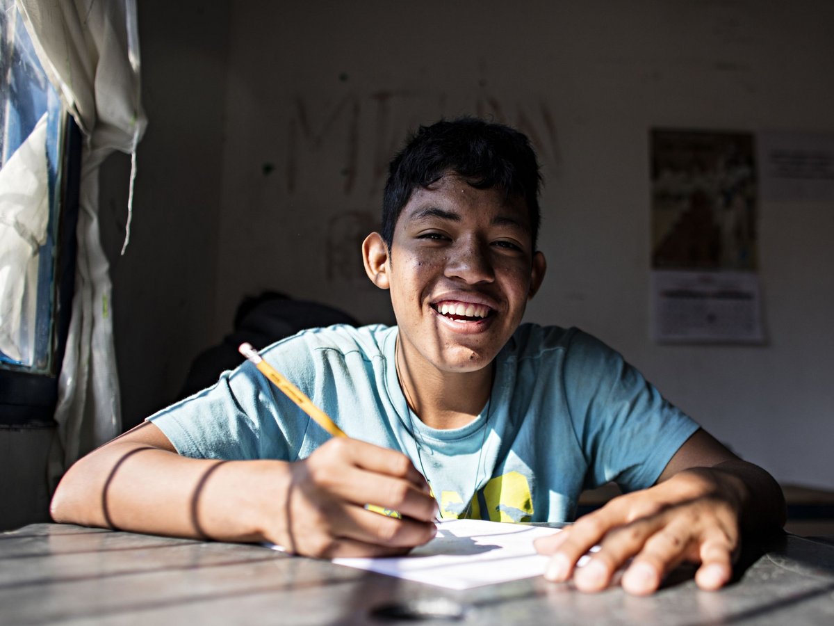 Lächelnder junger Mann im blauen Hemd schreibt an einem Schreibtisch, von der Sonne beleuchtet.