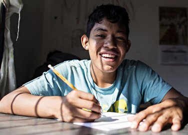 Lächelnder junger Mann im blauen Hemd schreibt an einem Schreibtisch, von der Sonne beleuchtet.