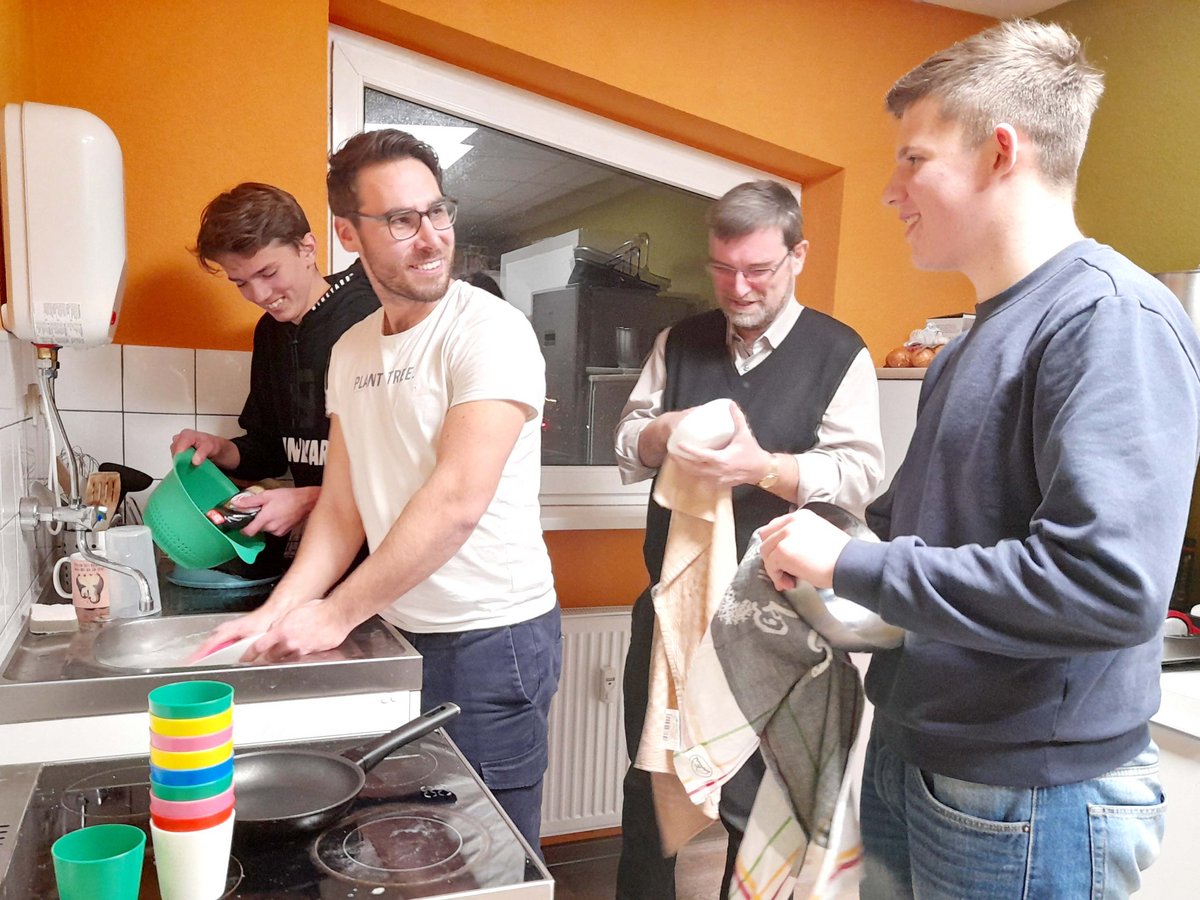 Vier Männer lächeln, während sie gemeinsam in einer Küche Geschirr spülen und abtrocknen.