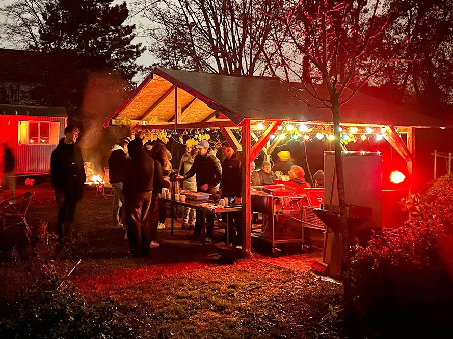 Nächtliches Gartenfest mit Leuten, die unter einem beleuchteten Pavillon feiern, umgeben von Lagerfeuer und rotem Licht.