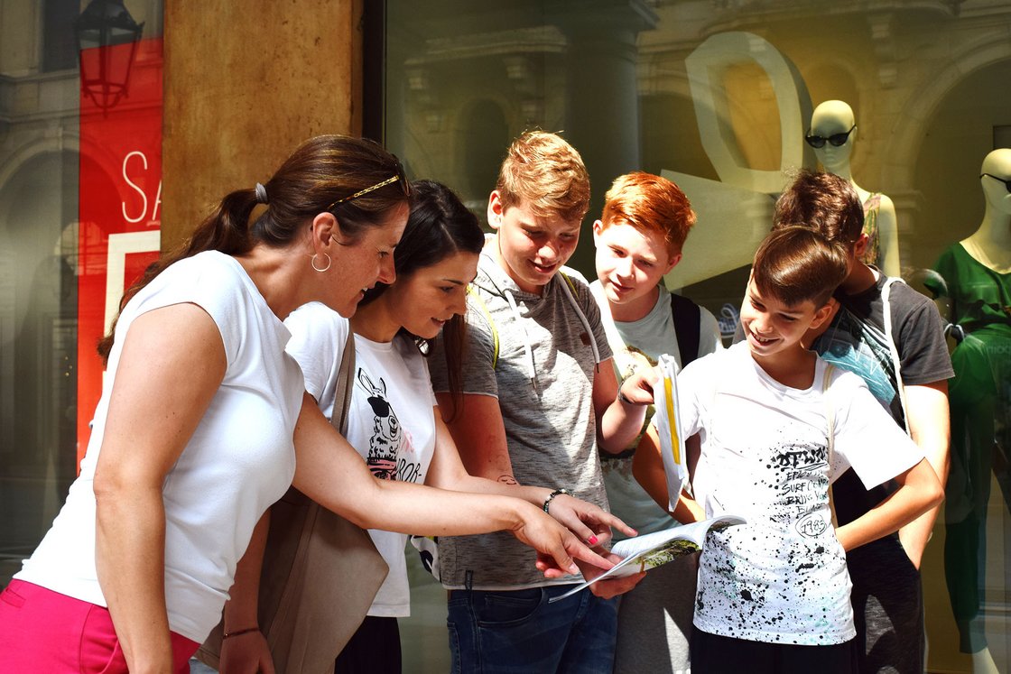 Eine Frau zeigt einer Gruppe von Jugendlichen und einer anderen Frau etwas in einem Reiseführer oder einer Karte auf der Straße.