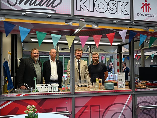 Vier Männer hinter einem geschmückten Kiosk-Tresen mit Getränken. Schilder für 'Smart KIOSK' und 'Don Bosco Jugend Nürnberg'.