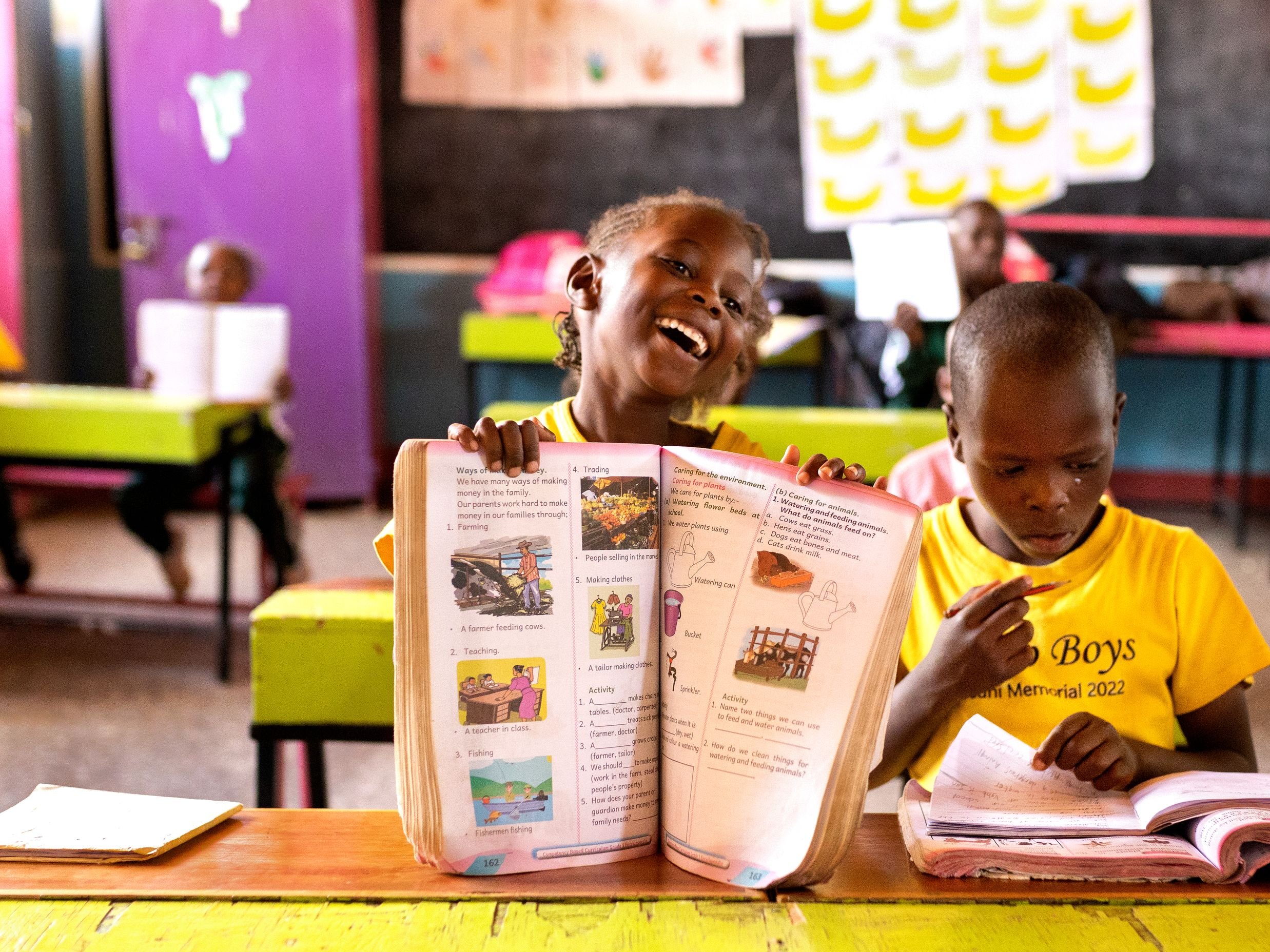 Ein fröhliches afrikanisches Mädchen lacht mit einem Lehrbuch, neben einem Jungen, der in einem farbigen Klassenzimmer schreibt.