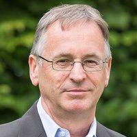 Porträt eines Mannes mit grauem Haar, Brille, Anzugjacke und einer kleinen Kreuznadel vor grünem Hintergrund.