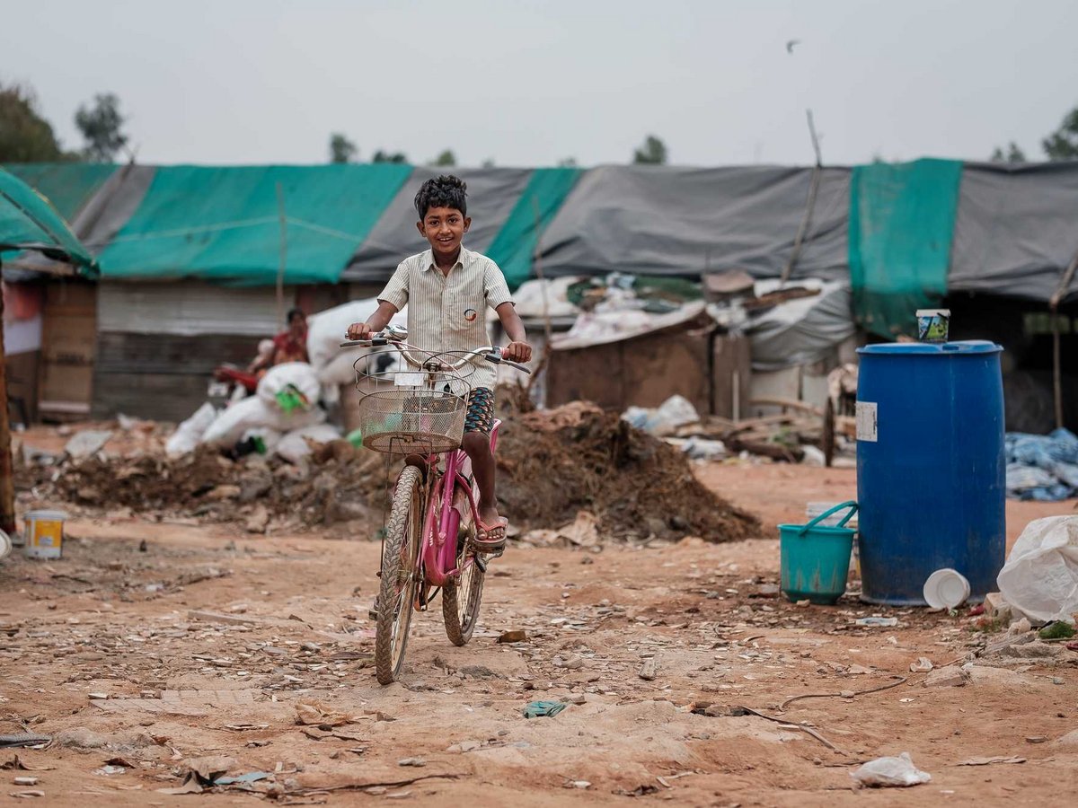 Junger Junge mit lockigem Haar lächelt beim Fahrradfahren auf einem Feldweg in einem Lager mit Behelfsunterkünften.