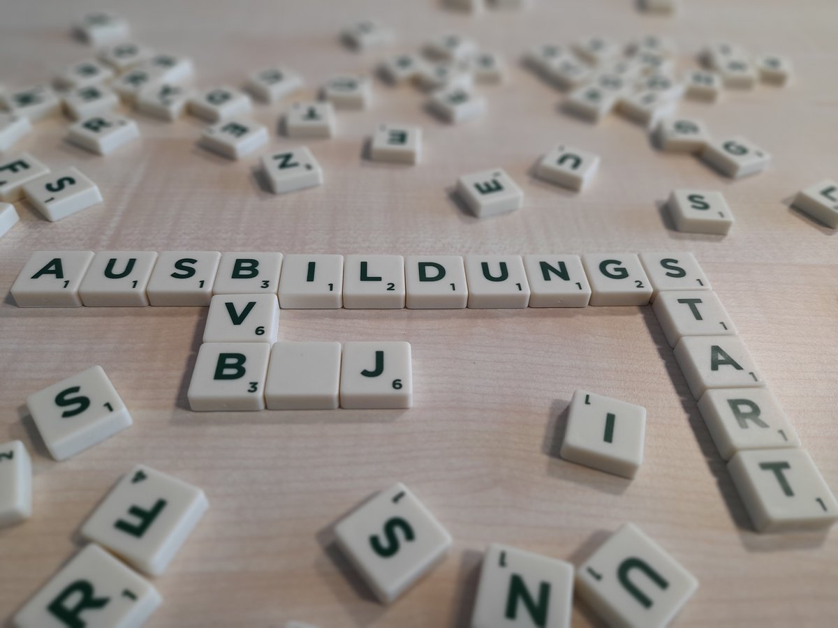 Scrabble-Steine bilden das Wort 'AUSBILDUNGSSTART' auf hellem Holzuntergrund, mit weiteren verstreuten Steinen.