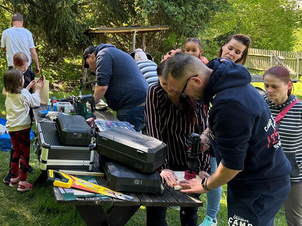 Eine Gruppe von Erwachsenen und Kindern bastelt draußen; ein Mann bohrt Holz an einem Tisch voller Werkzeuge.