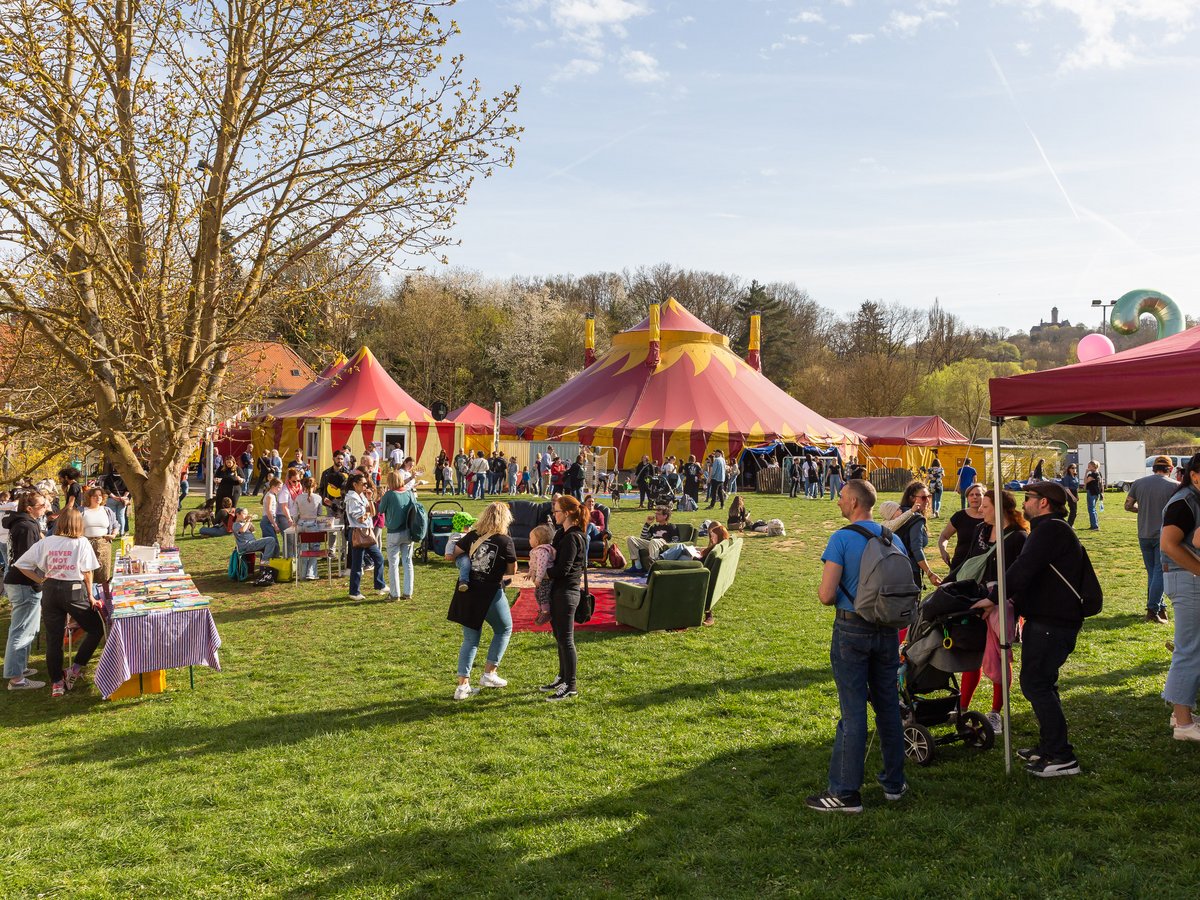 Menschen versammeln sich an einem sonnigen Tag auf einem Festival mit rot-gelben Zirkuszelten, einem Buchstand und einer Burg im Hintergrund.