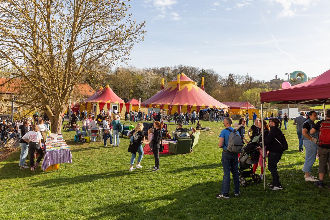 Menschen versammeln sich an einem sonnigen Tag auf einem Festival mit rot-gelben Zirkuszelten, einem Buchstand und einer Burg im Hintergrund.
