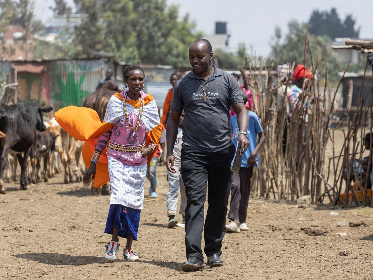 Eine traditionell gekleidete Frau und ein Mann in einem Poloshirt gehen auf einem sonnigen Feldweg vorbei an Kühen und einfachen Hütten.