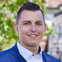 Lächelnder Mann in blauem Anzug und weißem Hemd, mit roter Anstecknadel, vor verschwommenem Außenhintergrund.