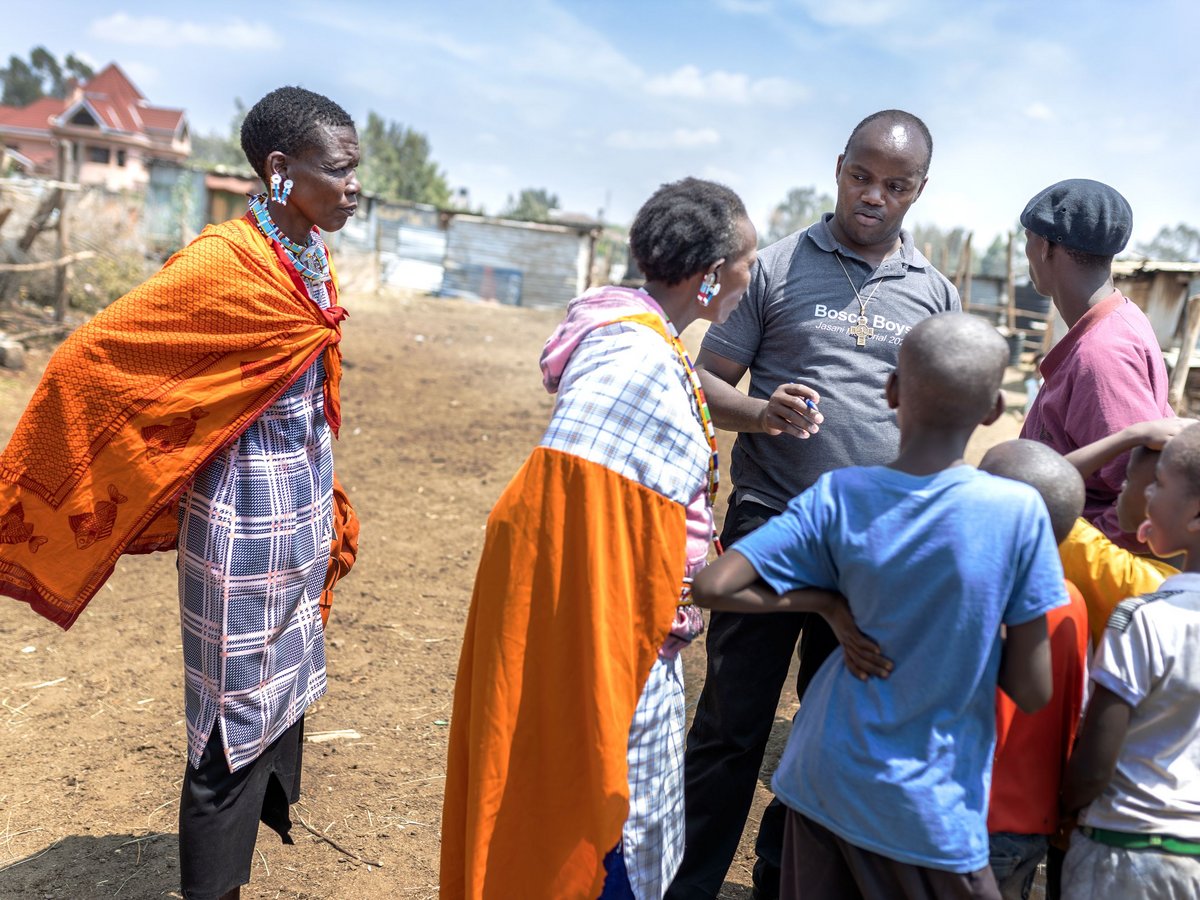 Ein Mann spricht mit Frauen in traditioneller Kleidung und Kindern im Freien bei Sonnenschein.