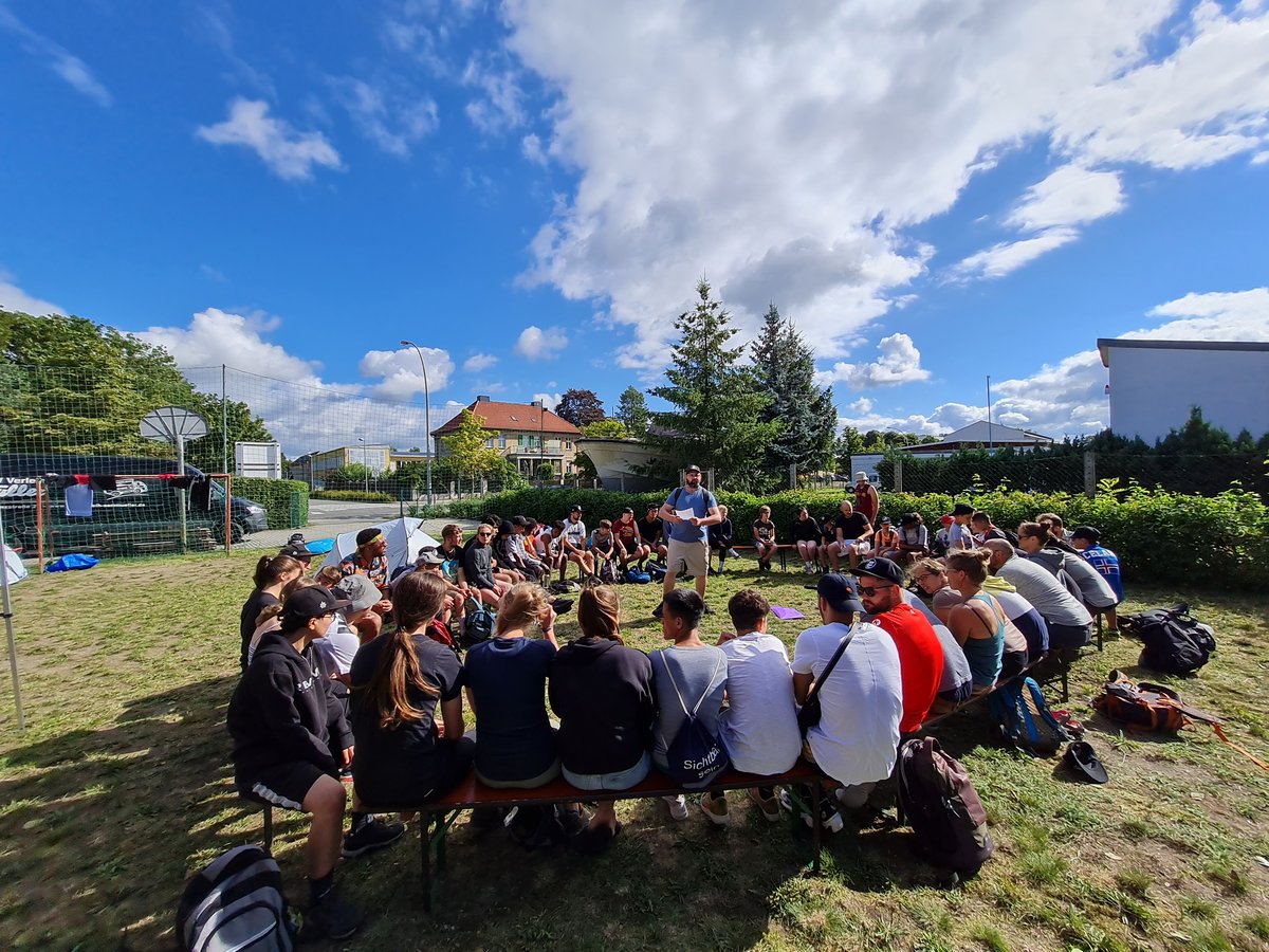 Jugendliche sitzen im Freien im Kreis auf Bänken, Betreuer steht in der Mitte und spricht Morgenimpuls