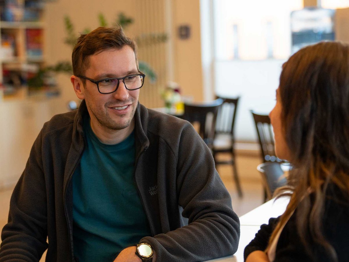 Ein lächelnder Mann mit Brille und Fleecejacke spricht mit einer Frau an einem Tisch in einem hellen Raum.