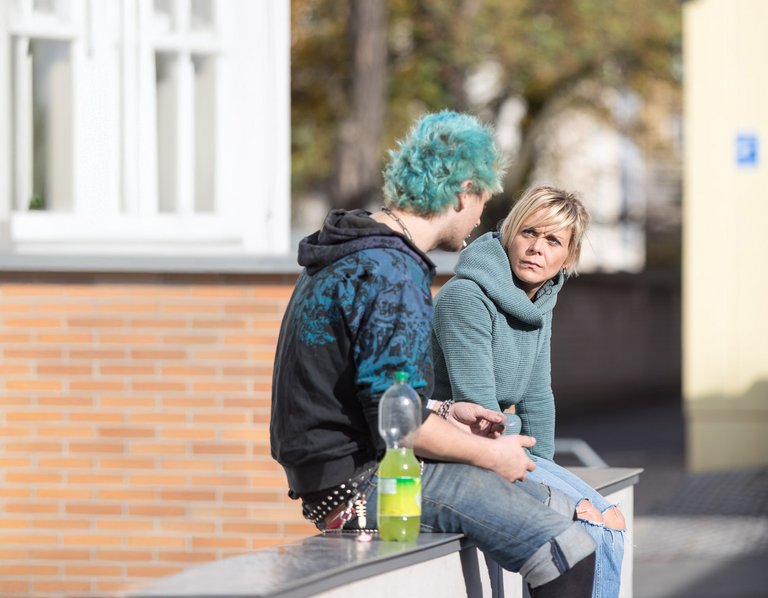 Ein Mann mit hellblauen Haaren und eine blonde Frau mit ernstem Ausdruck sitzen draußen, eine Flasche dazwischen.