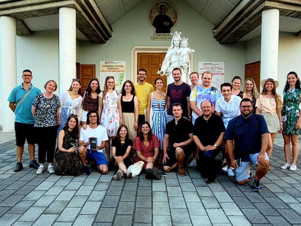 Lachende Gruppe junger und älterer Erwachsener posiert vor einer Kirche mit Marienstatue und heiligem Wandbild.