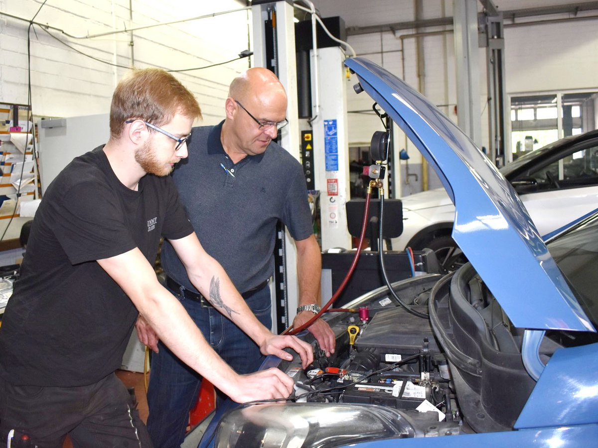Zwei Mechaniker inspizieren den Motor eines blauen Autos mit offener Motorhaube in einer Werkstatt. Diagnosegerät angeschlossen.