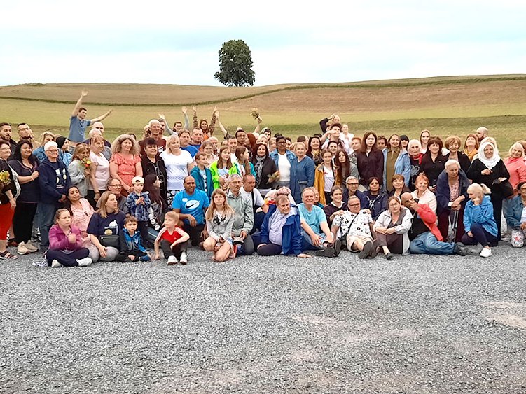 Eine große, vielfältige Gruppe von Menschen jeden Alters lächelt und posiert draußen auf einem Schotterweg mit Hügeln und einem Baum.