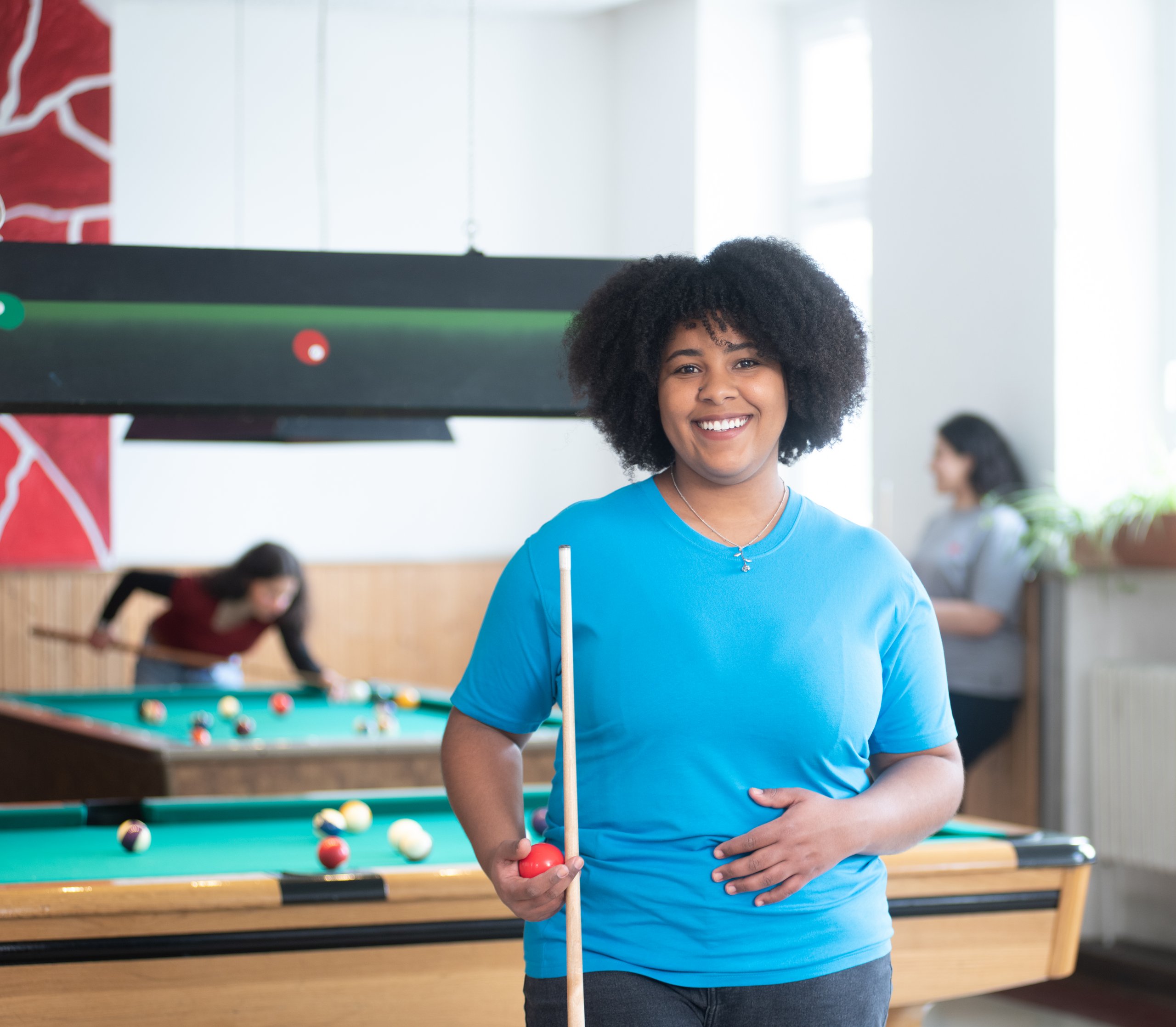 Lächelnde Person mit dunklem Lockenhaar hält Billardqueue und rote Kugel in einem Raum, in dem andere Billard spielen.