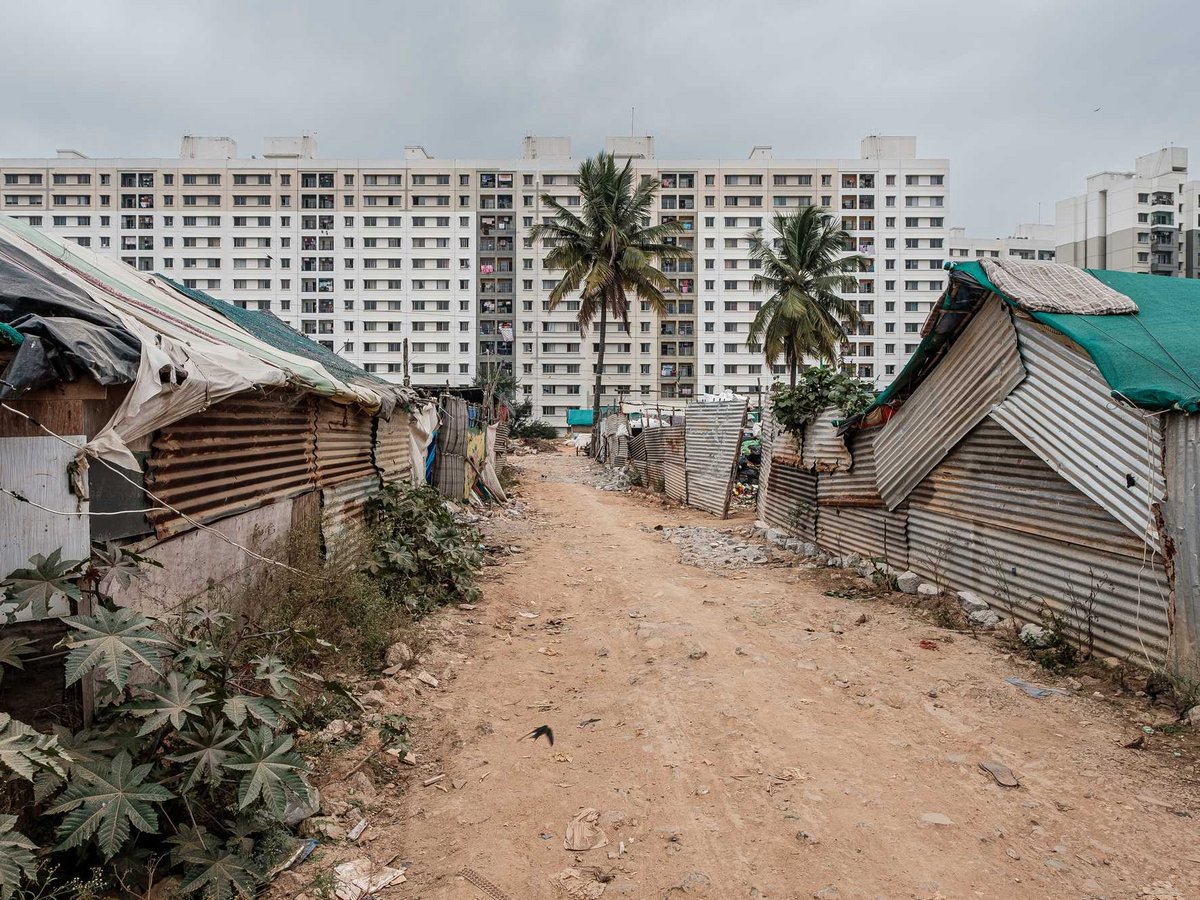 Hütten aus Metall und Planen säumen einen Feldweg, kontrastierend mit großen weißen Wohngebäuden und Palmen dahinter.