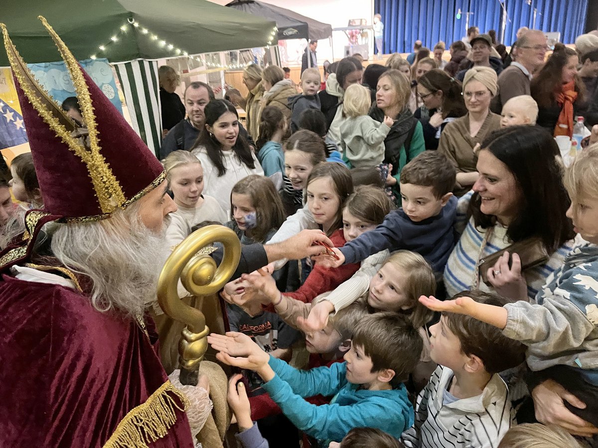 St. Nikolaus mit Bischofsmütze und Stab umringt von begeisterten Kindern, die die Hände nach Geschenken ausstrecken.