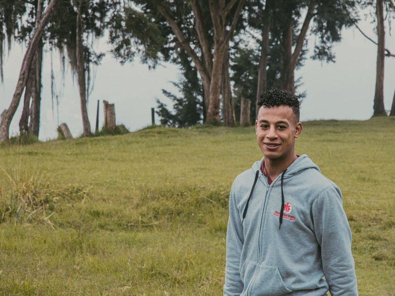 Ein lächelnder junger Mann mit lockigem Haar und Zahnspange in einem grauen Hoodie auf einem grasbewachsenen Hügel.