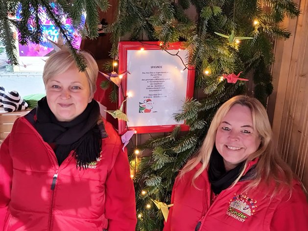 Zwei lächelnde Frauen in roten Jacken an einem festlichen Marktstand, geschmückt mit Tannengrün, Lichtern und Origami.