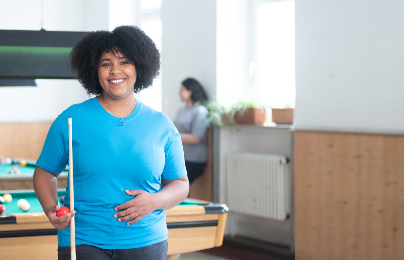 Lächelnde junge Frau mit Locken, Billardqueue und roter Kugel an einem Billardtisch.