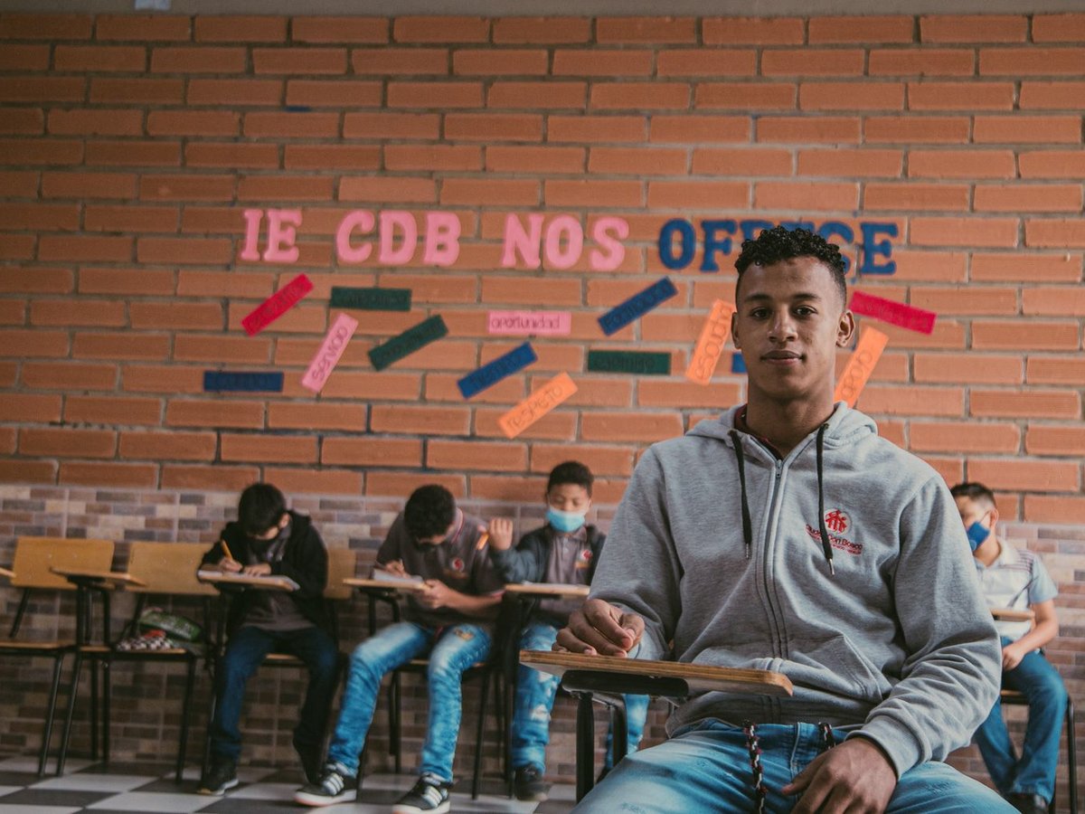 Junger Mann in grauem Kapuzenpullover sitzt in einem Klassenzimmer. Andere Schüler lernen im Hintergrund. Ziegelwand mit inspirierenden Wörtern.