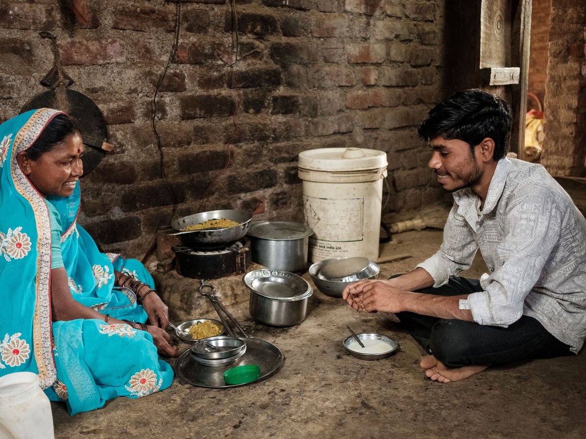 Lächelnde indische Frau im blauen Sari und junger Mann sitzen auf dem Boden, umgeben von Essen und Kochtöpfen.