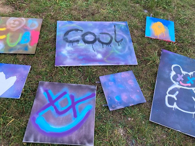 Verschiedene sprühbemalte Graffiti-Kunstwerke auf Leinwänden, mit Wörtern, Symbolen und einem Teddybär, auf Gras liegend.