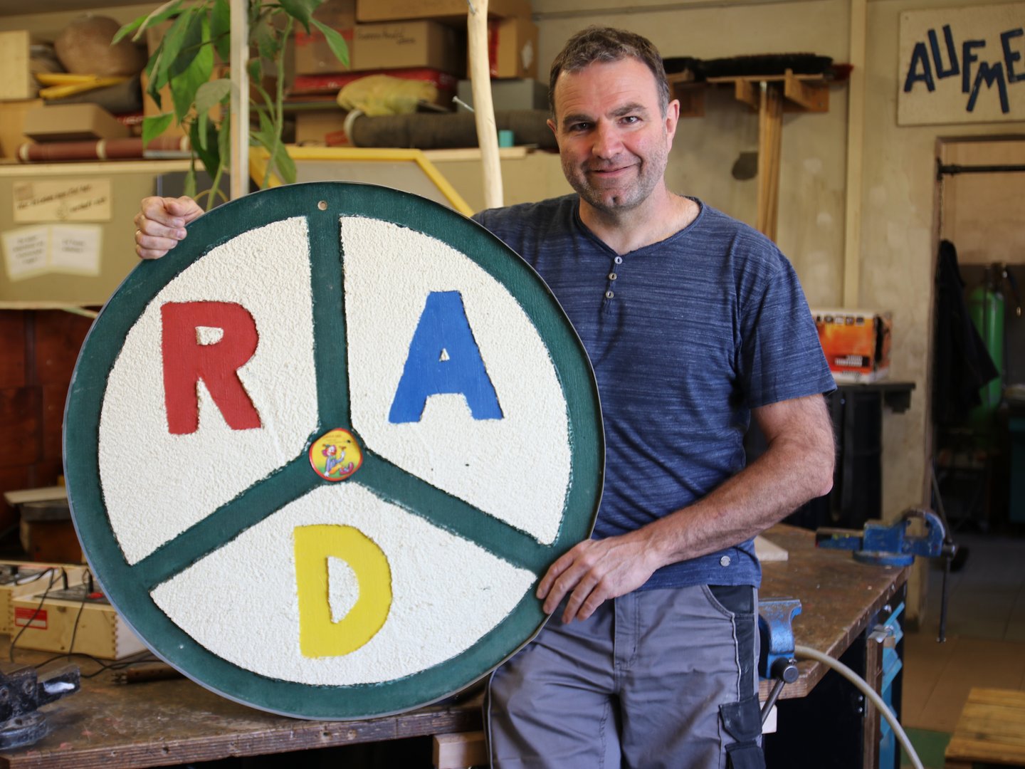 Ein lächelnder Mann in einer Werkstatt hält ein großes rundes Schild mit roten 'R', blauen 'A' und gelben 'D' Buchstaben, das einem Rad ähnelt.