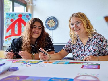Zwei junge Frauen lächeln in die Kamera, während sie an einem farbenfrohen Kunstprojekt mit Markern an einem Tisch arbeiten.